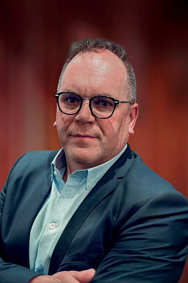 Portrait d’un homme d’affaires sérieux en costume sombre et chemise claire, portant des lunettes et regardant l’objectif avec un fond rouge flouté.