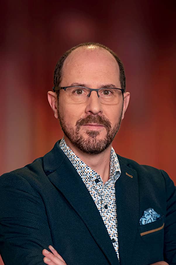 Portrait d’un homme aux lunettes et barbe soignée, vêtu d’une veste bleu foncé et d’une chemise à motifs, posé devant un fond flou aux tons chauds.
