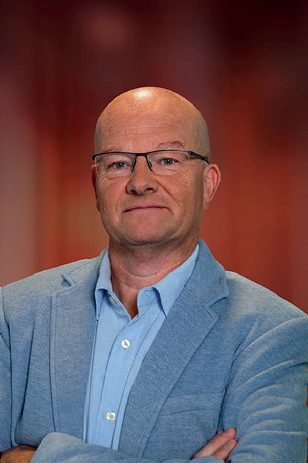 Homme d’affaires chauve portant des lunettes, veste bleu clair et chemise, bras croisés devant un fond flou aux tons chauds