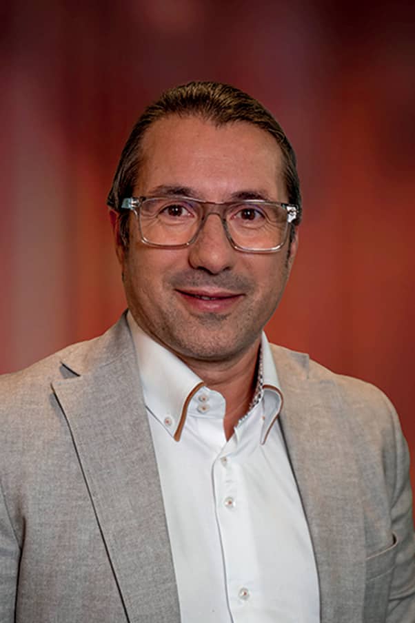 Homme d’âge moyen portant des lunettes transparentes, une chemise blanche et un blazer gris clair, souriant devant un fond rouge flou.