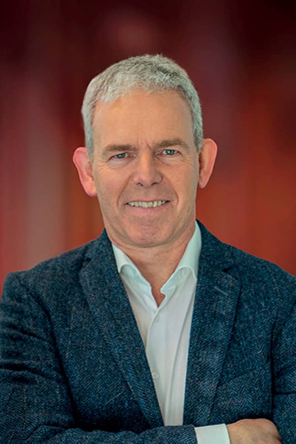 Portrait d'un homme d'affaires à la quarantaine aux cheveux grisonnants, souriant, bras croisés dans un blazer bleu foncé et chemise blanche sur fond rouge uni.