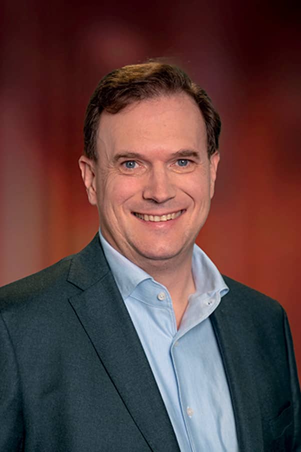 Portrait en buste d’un professionnel souriant vêtu d’un blazer foncé et d’une chemise bleu clair sur un fond flou aux teintes chaudes