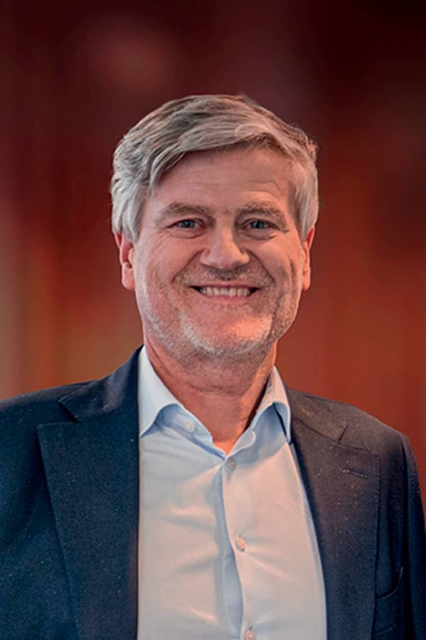 Portrait d’un homme d’affaires d’âge mûr avec cheveux gris, souriant, portant un costume bleu marine et une chemise blanche, sur fond flou aux tons chauds.