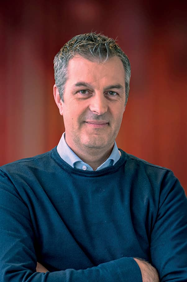 Homme d'environ 40 ans aux cheveux courts poivre et sel, portant un sweat-shirt bleu marine sur une chemise gris clair, les bras croisés et regardant l'objectif devant un fond flou rouge-brun.