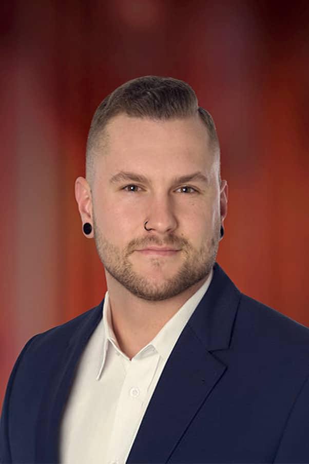 Portrait d’un jeune homme barbu, avec piercing nasal et boucles d’oreilles, portant une chemise blanche et un veston bleu foncé devant un fond flou rouge-orangé