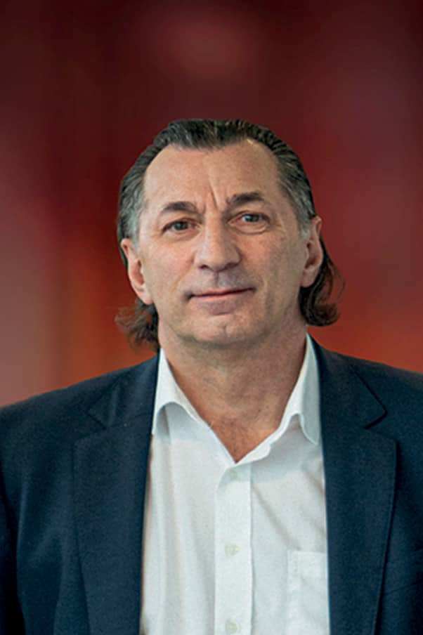 Portrait en buste d’un homme d’âge moyen portant une chemise blanche et un blazer bleu devant un fond rouge flou