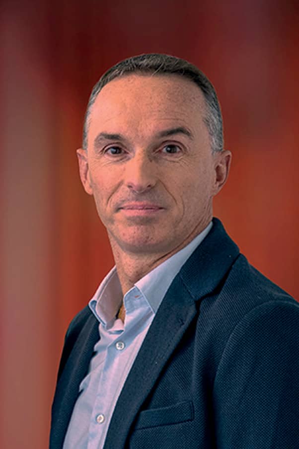 Portrait d’un homme d’âge moyen portant un blazer bleu marine et une chemise bleu clair, au regard confiant, sur un fond orange flouté à lignes verticales.
