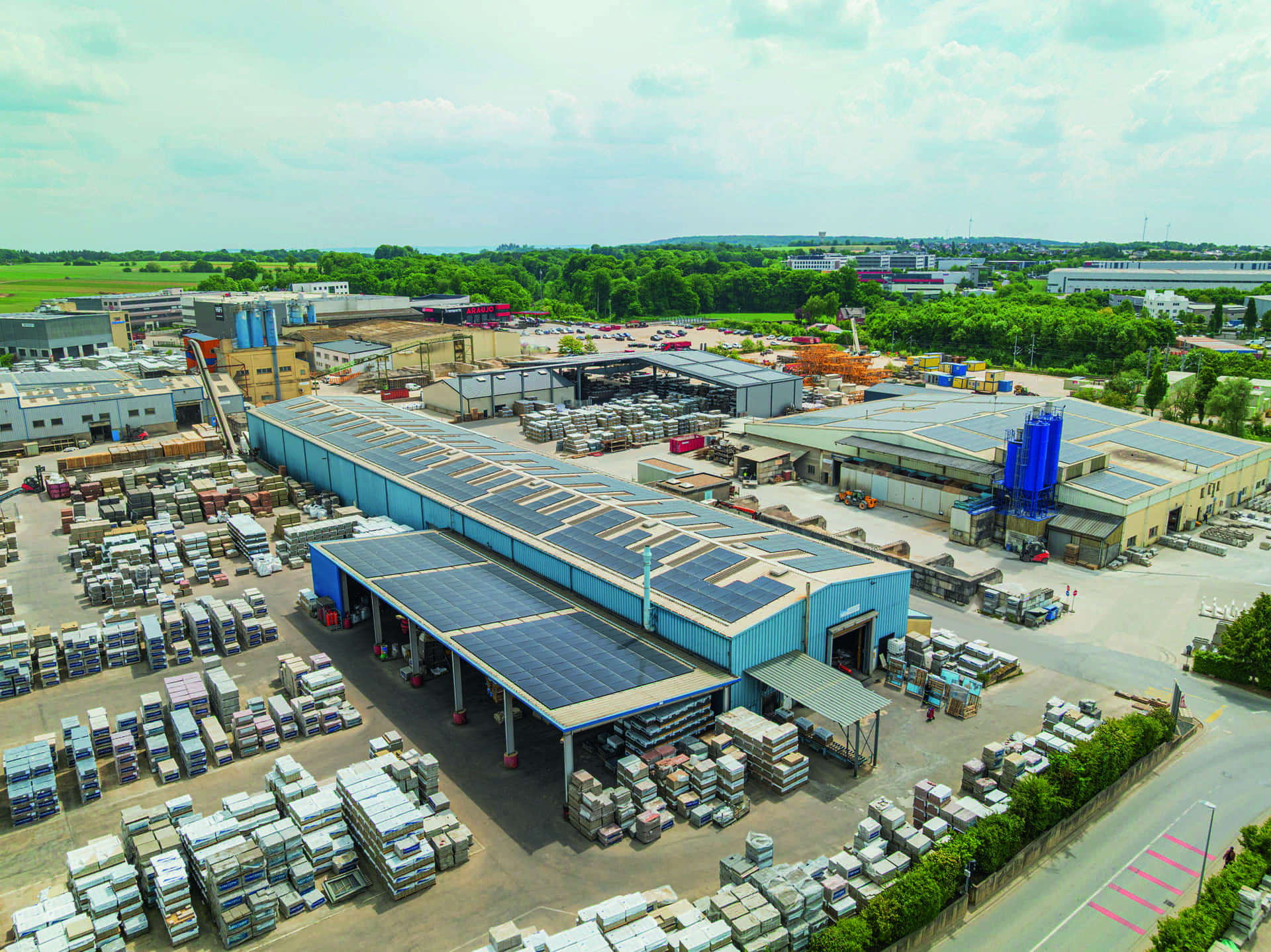 Vue aérienne d’un parc industriel de stockage de pierres présentant des centaines de dalles et blocs empilés sur palettes autour de grandes halles bleues couvertes de panneaux solaires, un parking, des champs verts et un bâtiment marqué « ARAUJO »